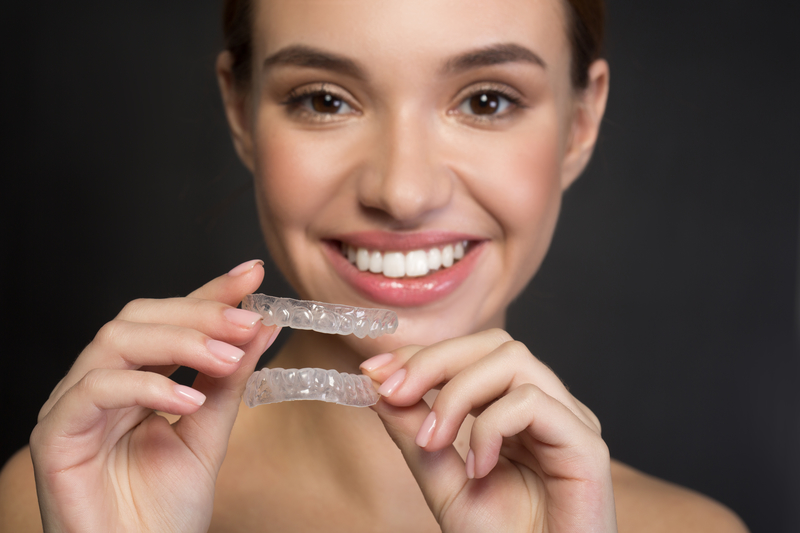 A beautiful brunette woman that is smiling and holding transparent orthodontic aligners in front of her.