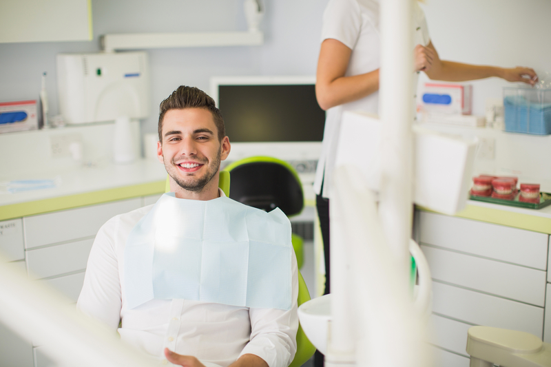 An attractive, young adult male that is sitting in a dental chair as a dental hygienist prepares tools in the background.